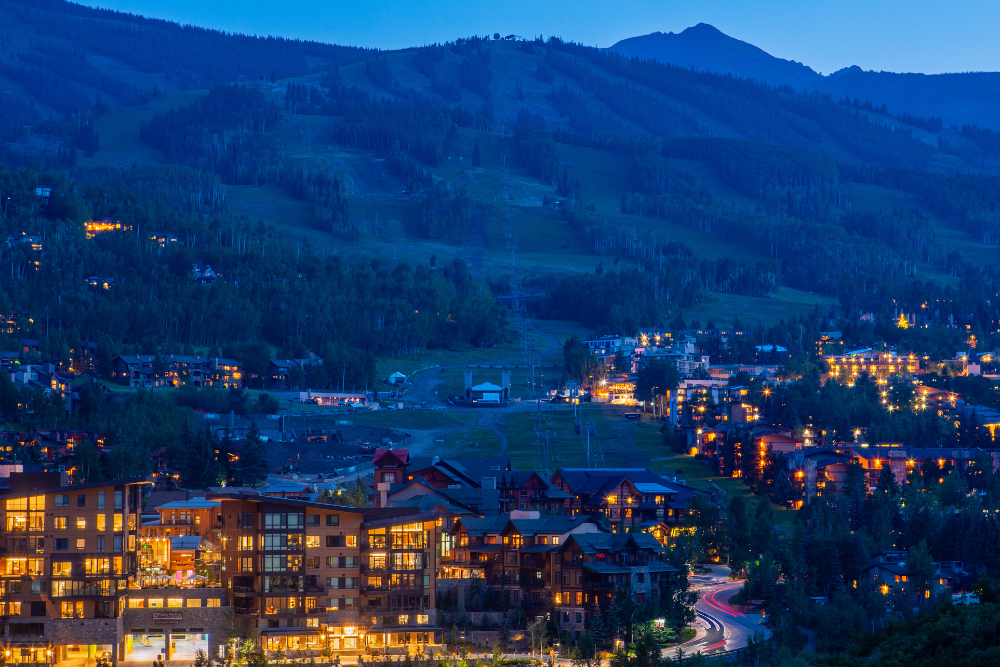 A hillside mountain village at dusk with glowing windows and streetlights along winding roads, set against dark green peaks under a blue sky.