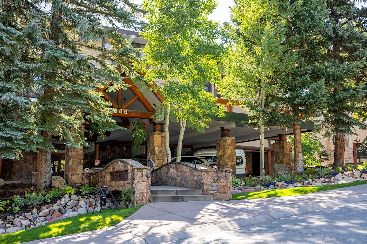 Lush trees frame a building entrance with stone walls, landscaped grounds, and parked cars beneath a green awning.