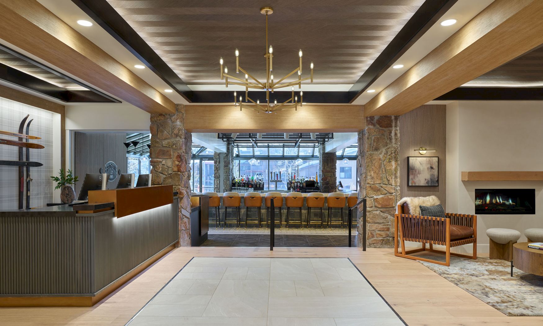 Modern lounge/bar with stone columns, gold chandelier, wooden stools by the counter, and a lit fireplace in warm light.