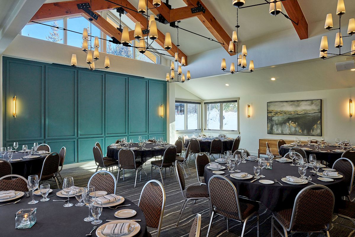 An elegant dining room with set tables, warm chandeliers, dark wood beams, and teal paneled walls under skylit windows.