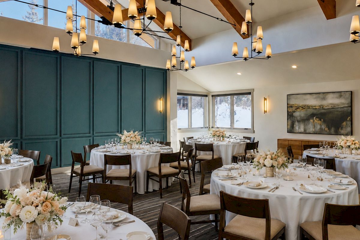 An elegant banquet room set for a wedding or dinner, with chandeliers, greenery wall panels, and floral centerpiece tables.