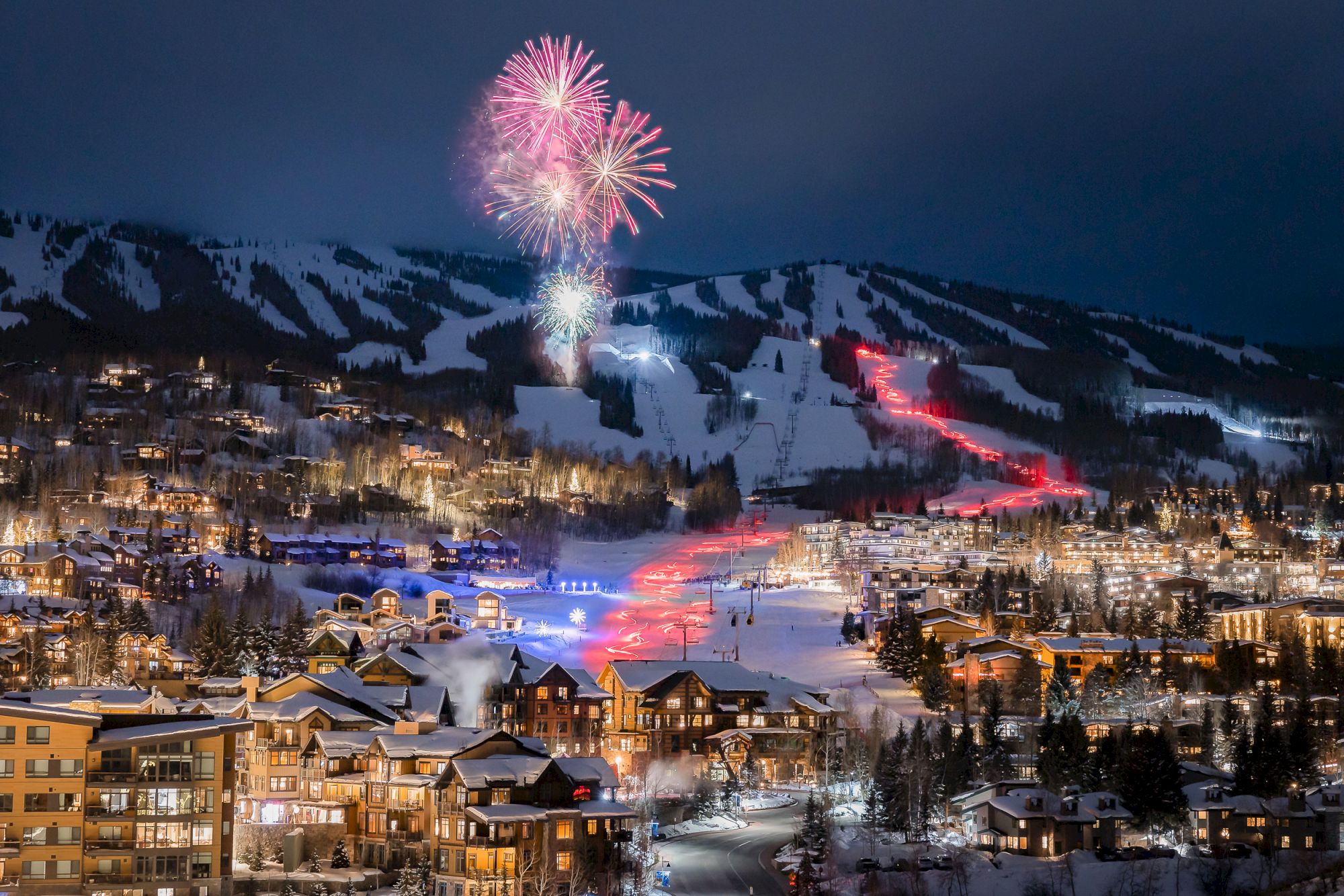 Fireworks burst over a snowy mountain ski town at night, while glowing lights and a winding road snake through the valley.