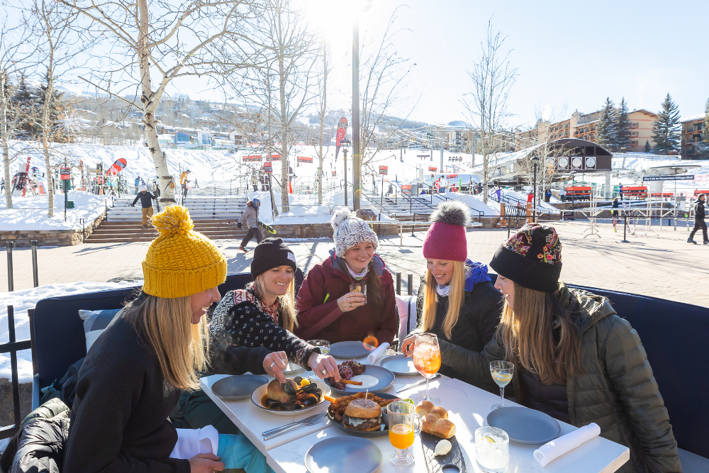 Five friends in winter hats share brunch outdoors at a patio table, with snow and a construction site in the background.