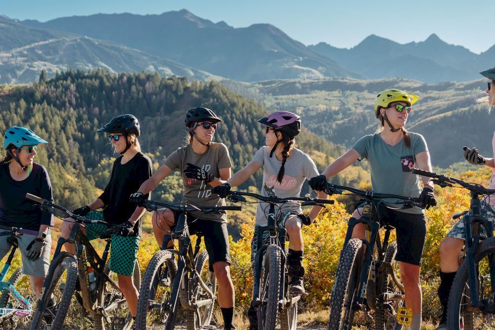 Helmed mountain bikers pause with their bikes on a trail, autumn hills and towering mountains behind them.