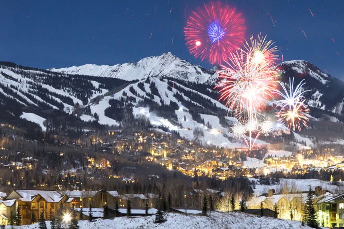 Fireworks burst over a snowy mountain village at night, with glowing lights and dark peaks in the background.