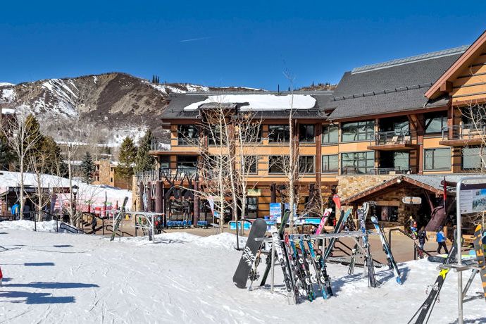 Snowy ski resort scene with skiers, a ski-school sign, and rows of rental skis beside buildings.