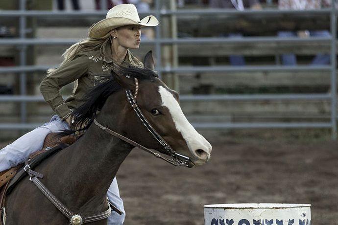 A cowgirl in a cowboy hat rides a dark-and-white horse in a rodeo arena near a barrel, captured mid-motion.