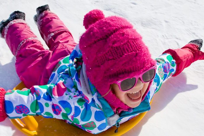 Two kids in bright winter suits and hats sled down snowy slopes, smiling in sunglasses while riding a colorful snow tube.