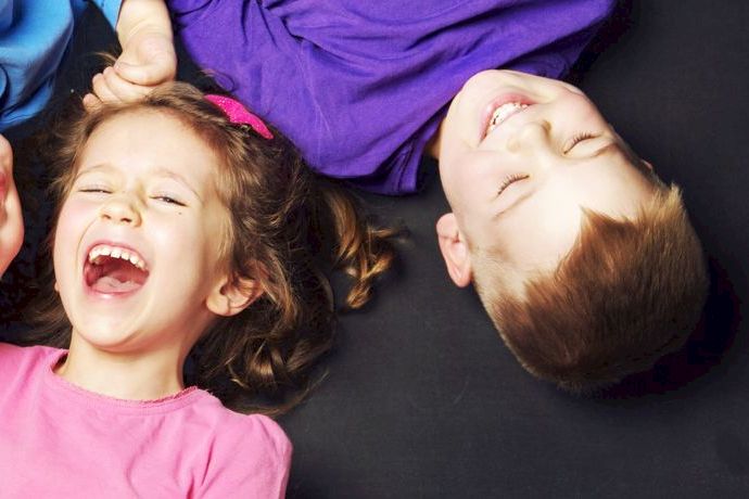 Four kids lie upside down, laughing together on a dark background, their faces bright with joy.