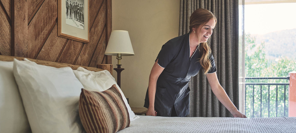 A woman in a hotel room adjusts a neatly made bed, smiling by the window with curtains and framed wall decor.