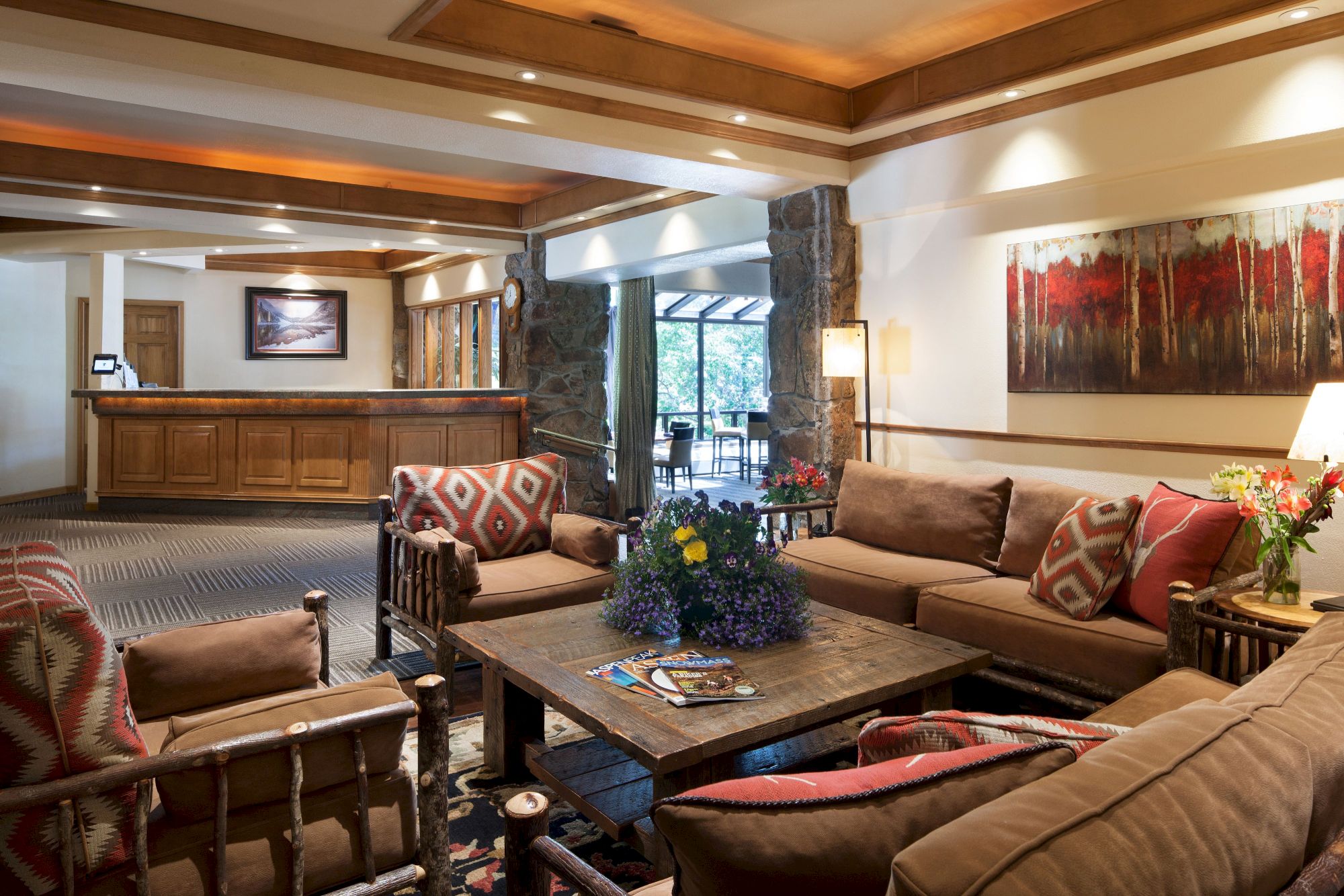 A cozy lodge-style living room with stone fireplace, wood beams, comfy sofas, colorful pillows, and a table in warm lighting.