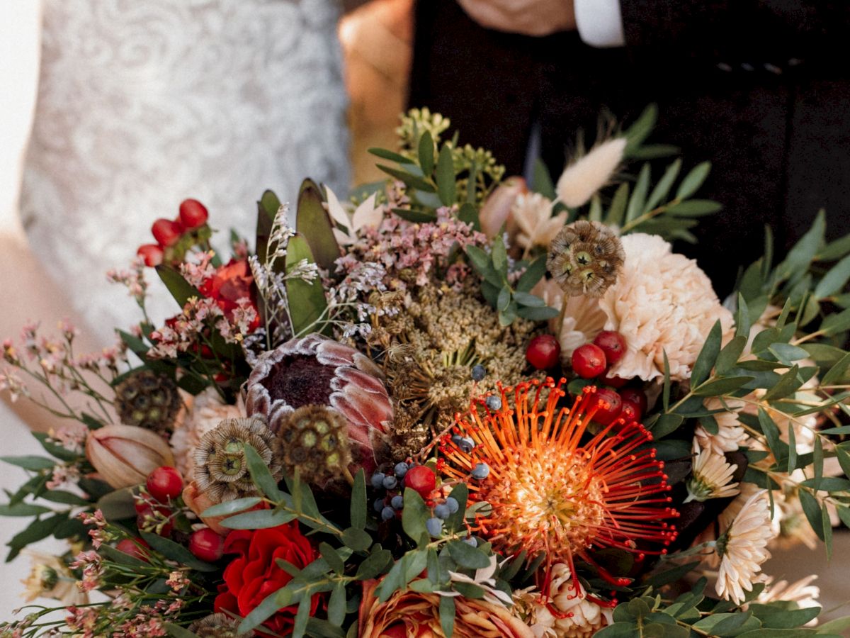 A large mixed flower bouquet with red and orange blooms, greenery, berries, and pinecone accents in someone&rsquo;s hands.