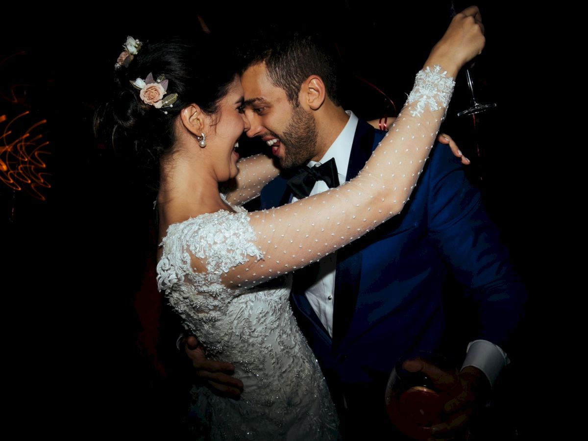 A bride and groom in formal wear dance and laugh at night, the groom&rsquo;s arm around her in a romantic hug.