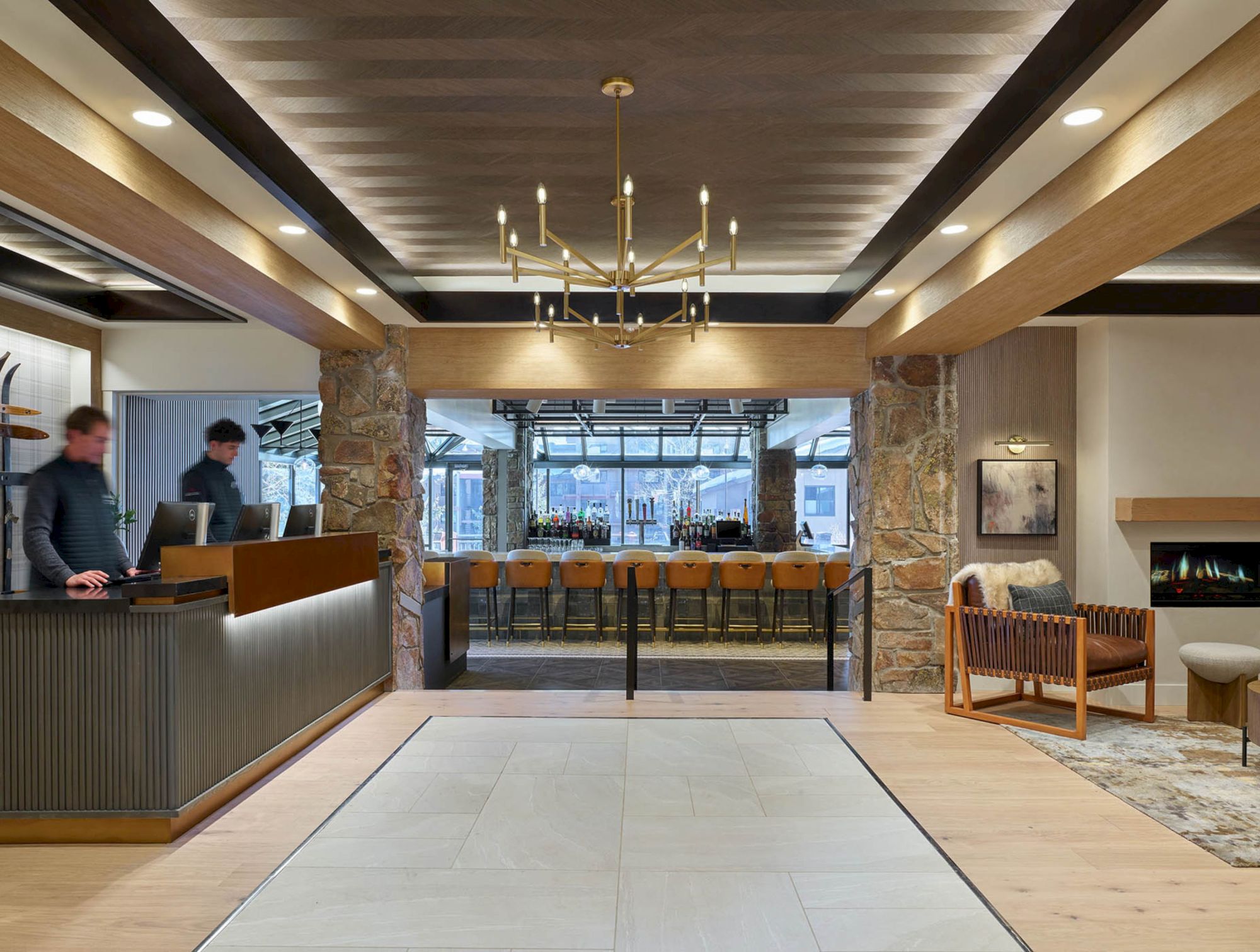 Modern bar interior with a gold chandelier, stone pillars, a bar counter, wine bottles, and yellow stools for guests.