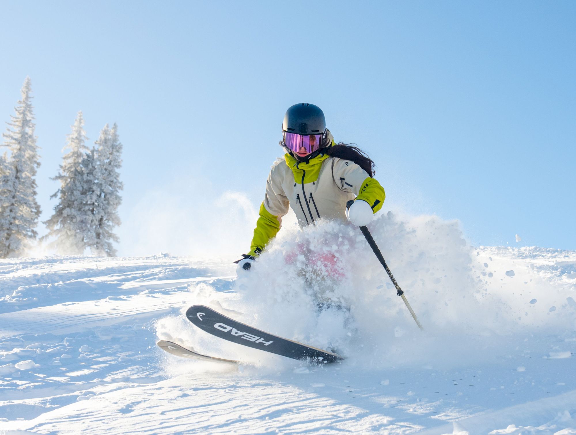 A skier in a helmet and goggles carving through fresh snow, with icy pines in the background under a clear blue sky.