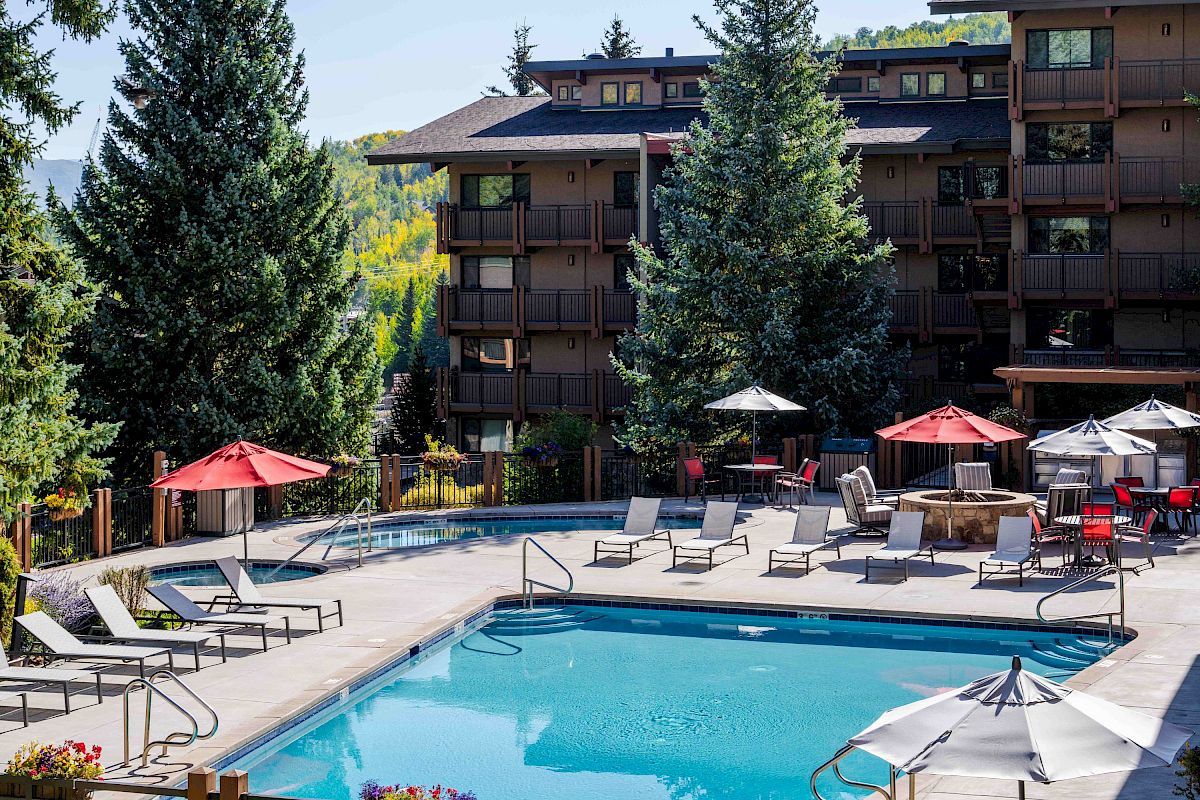 Sunlit hotel courtyard with a swimming pool, lounge chairs, umbrellas, and pine trees before modern multi-story buildings.