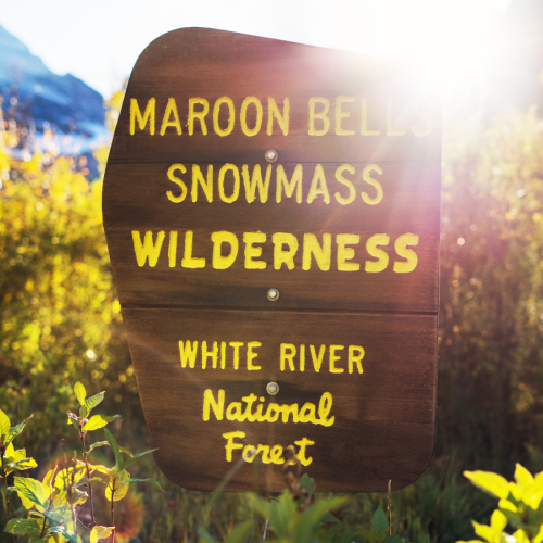 A wooden trail sign reads &ldquo;Maron Bell, Snowmass, Wilderness, White River National Forest&rdquo; in yellow letters.