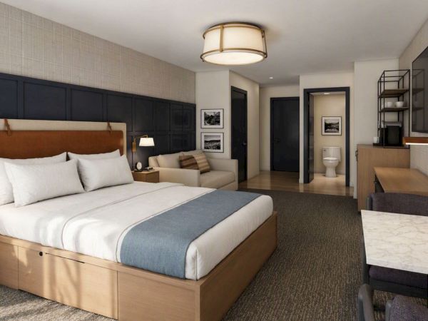 A modern hotel suite: made bed, lamps, wall paneling, and an adjoining bathroom with vanity, mirror, and toilet.