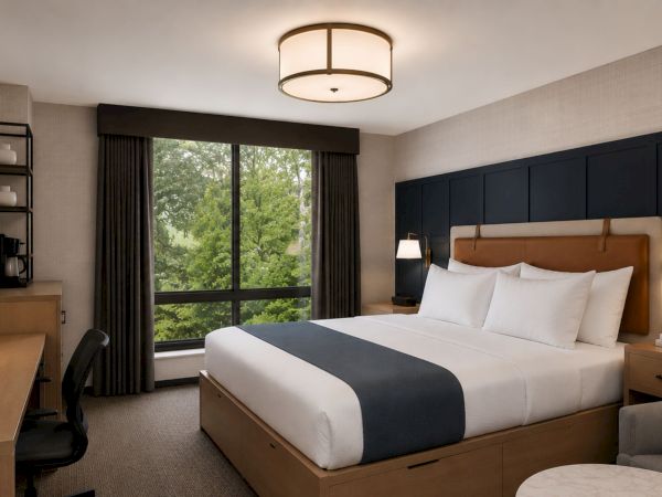 A modern hotel bedroom with a neatly made bed, bedside lamps, a large window, curtains, and a desk with chair.