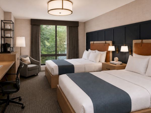 A modern hotel room with twin beds, dark headboard, desk and chair, coffee setup, and a large window with curtains.