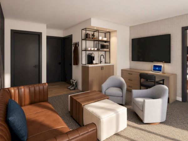 Modern hotel suite with leather sofa, chairs, coffee table, desk with laptop, dual TVs, and built-in kitchenette and wardrobe.