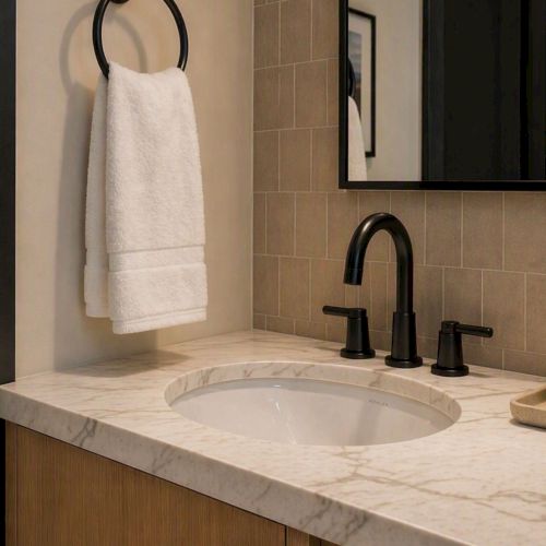 A modern bathroom vanity with a marble sink, black faucet, rolled towels, a mirror, and three bottles of toiletries on a tray.