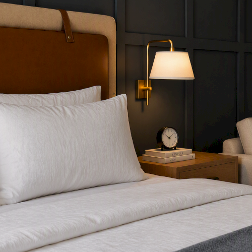 A modern bedroom with a bed, white pillows, a brass lamp, a clock and books on the nightstand, and wall art beside a sofa.