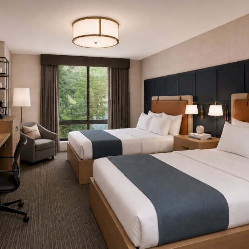 A modern hotel room with two beds, bedside lamps, a desk, and a window showing green trees.