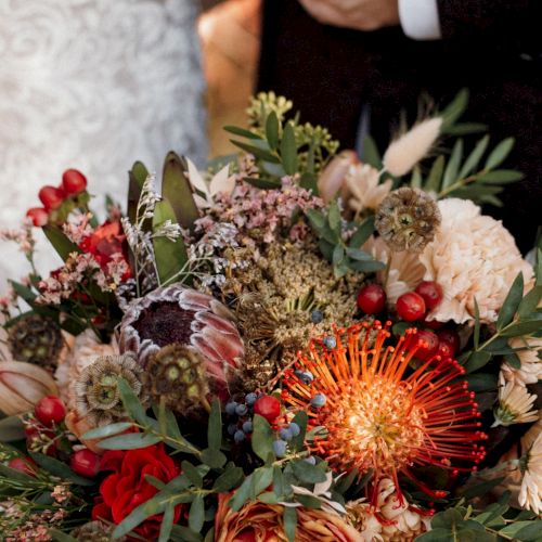 A large hand-held bouquet packed with orange pincushion flowers, red berries, cream blooms, eucalyptus leaves, and dried accents.