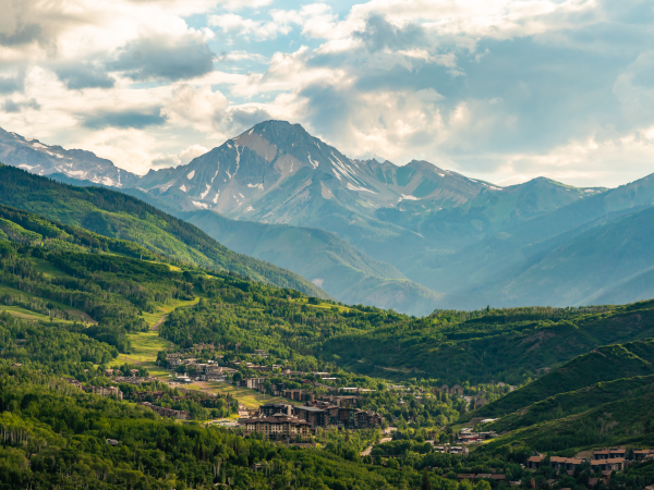 A scenic mountain valley with pine forests, a small town below, and dramatic peaks under a cloudy, sunlit sky.