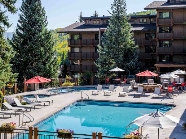 A sunny resort pool deck with lounge chairs, red/white umbrellas, and a multi-story apartment building surrounded by trees.
