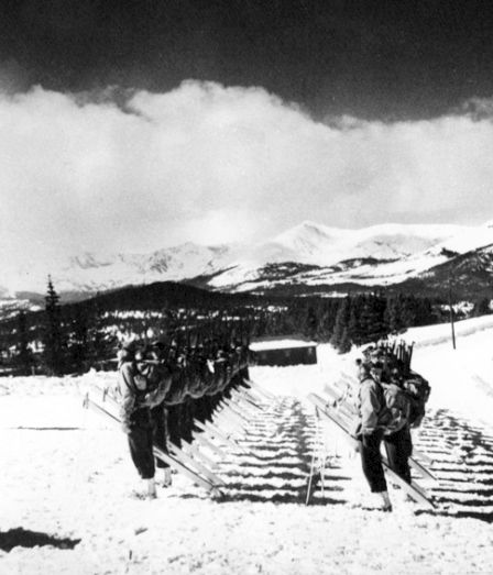 A line of soldiers with rifles marches through snowy countryside toward the mountains, surrounded by snow-covered trees.