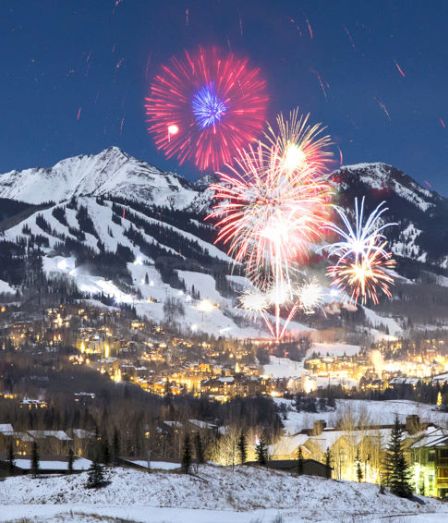 Fireworks burst over a snowy alpine village with glowing lights below and dark mountains under a blue night sky.