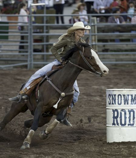 A cowgirl in a hat rides a dark horse through a dusty rodeo arena, passing a barrel labeled &ldquo;SNOWMAN RODE.&rdquo;