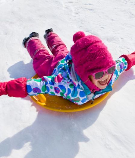 A child in bright pink winter clothes rides a yellow inner-tube sled down a snowy slope, laughing in the sunshine.