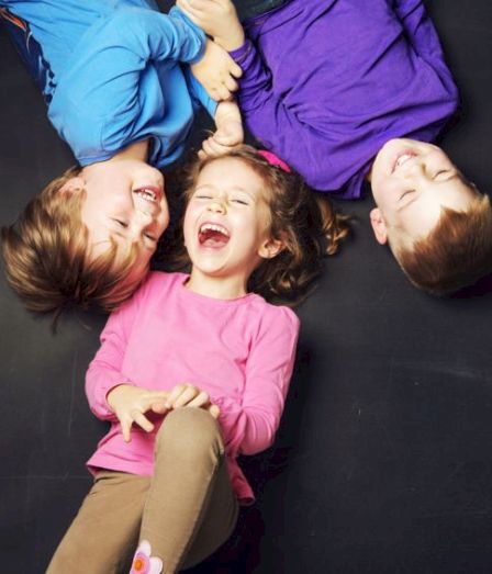 A bright photo of three kids lying on a dark floor, laughing and smiling as they play together.