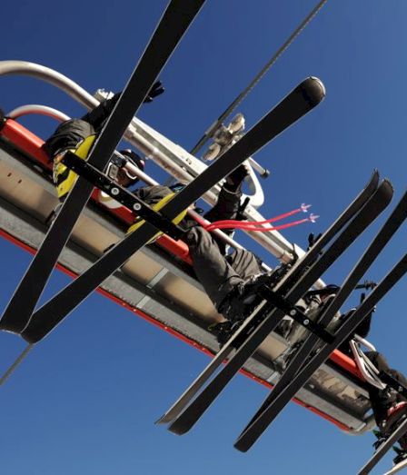 A skyward view of a worker secured in a harness on a large industrial mast with ropes and cables against a blue sky.