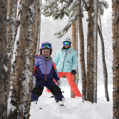 Two kids in colorful winter gear and goggles stand in deep snow between tall icy trees, smiling after a snowy adventure.