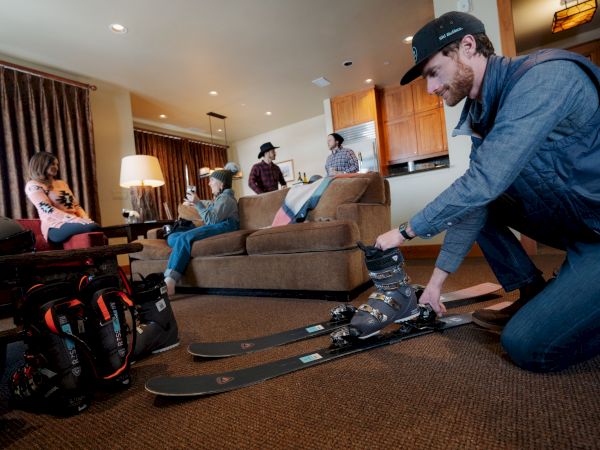 A man in a cap adjusts ski boots in a hotel living room while others watch, with skis and helmets nearby.
