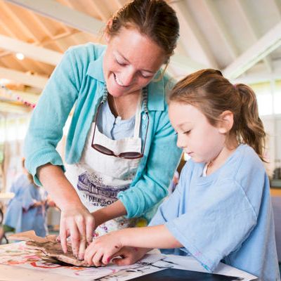 A craft instructor teaches a young child to draw and paint together at a colorful table indoors.