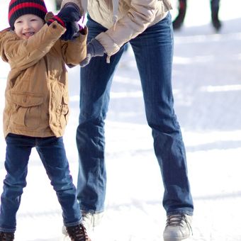 A child in a winter coat and beanie holds an adult&rsquo;s hand while standing in the snow.