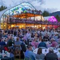 A packed outdoor festival audience dining and enjoying a live concert on a brightly lit stage under an arched canopy.