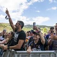 A cheering crowd presses against barricades at an outdoor concert with mountains in the background, with people waving and smiling.