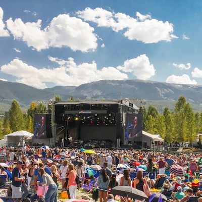 A packed outdoor music festival crowd cheers in front of a big stage, with green mountains and blue skies behind them.
