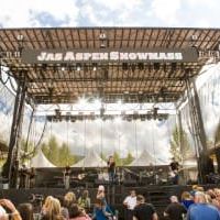 Outdoor music festival stage with a crowd, banners, and mountain-like backdrop under a sunny sky.
