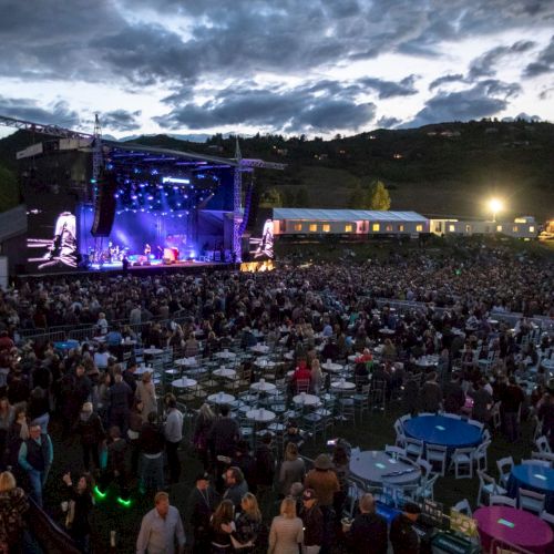 An outdoor music festival at dusk with a lit main stage and a huge crowd packed into stadium-style seating and tents.