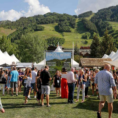People mingle at an outdoor Food & Wine festival in a grassy valley, with white tents and green mountains in the background.