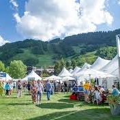 An outdoor food-and-wine festival with white tents, people mingling on grass, and green mountains in the background.