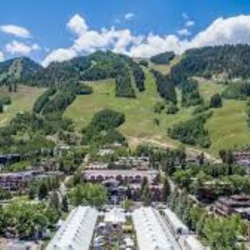 A lush alpine valley with evergreen hills, a small village, and snowy mountain peaks under a bright blue sky.