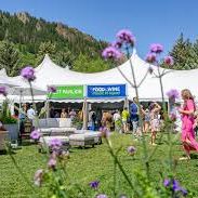 Outdoor market with vendors and shoppers in a mountain meadow, surrounded by colorful flowers and flags.